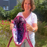 Woman holding a pink and purple patterned bag outdoors with greenery and flowers in the background