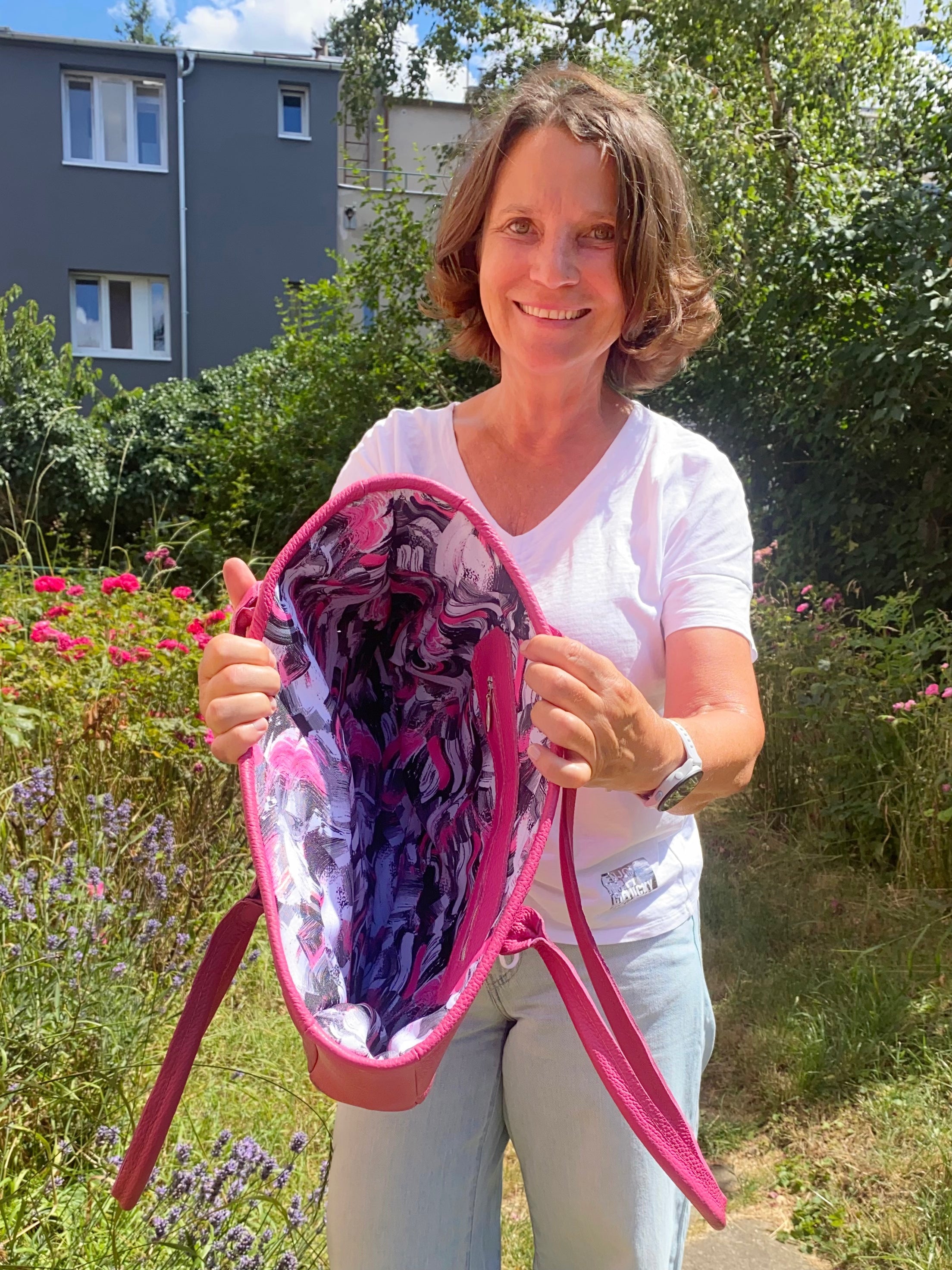 Woman holding a pink and purple patterned bag outdoors with greenery and flowers in the background