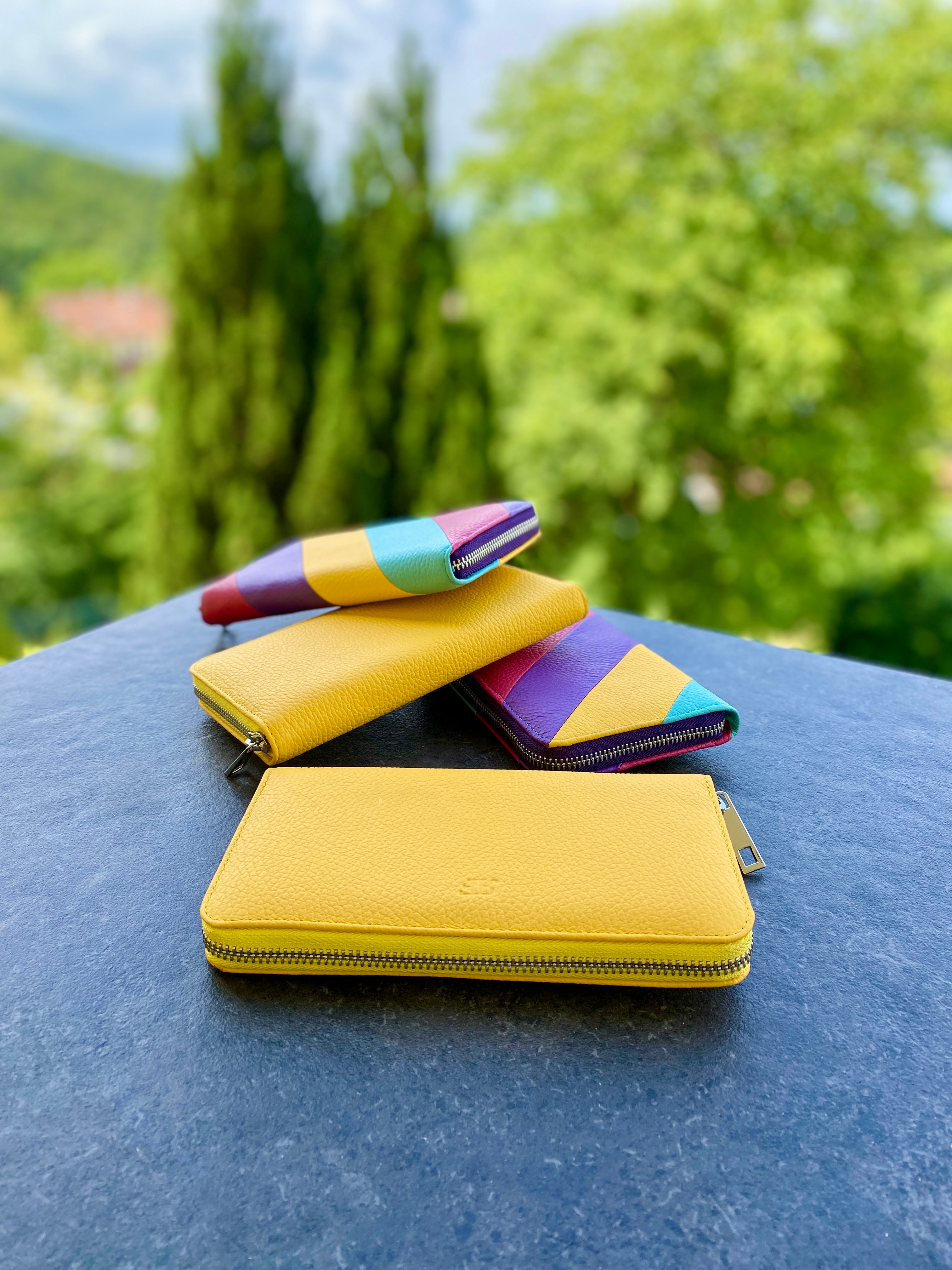 Yellow wallet, pink and purple pen, and pink and yellow wallet on a blue surface with greenery in the background