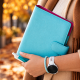 A person in a beige coat and brown scarf holds a bright turquoise blue faux-leather notebook cover with a magenta inner lining, set against a blurred autumn park background with fallen leaves.