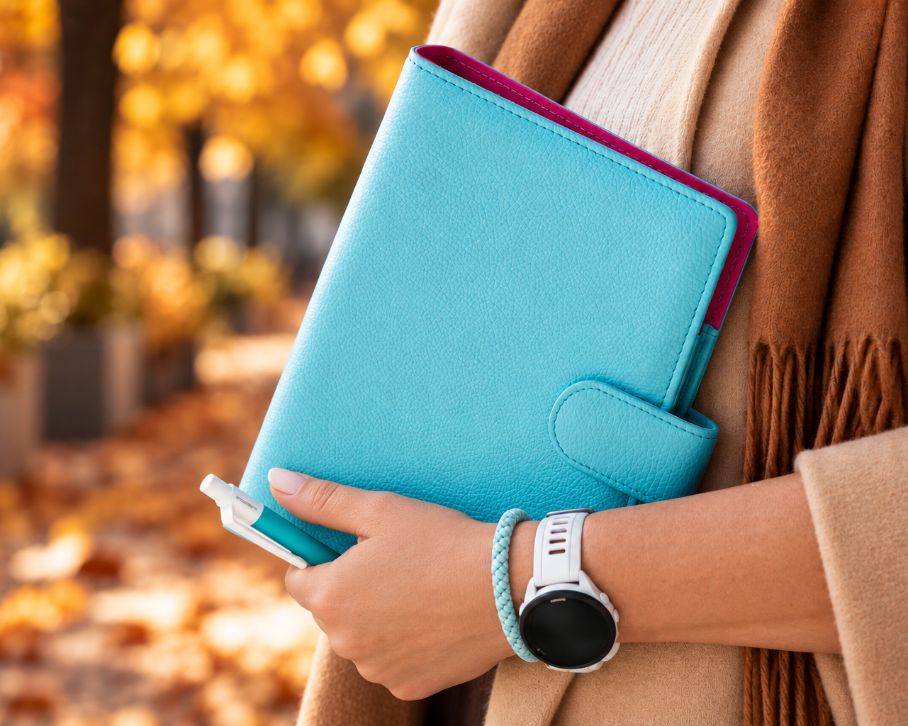 A person in a beige coat and brown scarf holds a bright turquoise blue faux-leather notebook cover with a magenta inner lining, set against a blurred autumn park background with fallen leaves.