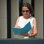 A woman wearing sunglasses and a sleeveless cream top stands behind a counter, holding a bright blue folder and writing inside it.
