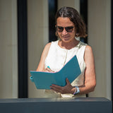 A woman wearing sunglasses and a sleeveless cream top stands behind a counter, holding a bright blue folder and writing inside it.