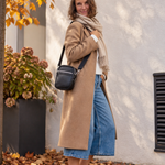 A woman stands outdoors on a stone walkway covered with fallen leaves, wearing a long camel coat, cream scarf, blue denim shorts, white sneakers, and carrying a black crossbody bag.