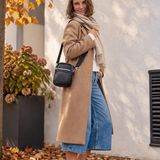 A woman stands outdoors on a stone walkway covered with fallen leaves, wearing a long camel coat, cream scarf, blue denim shorts, white sneakers, and carrying a black crossbody bag.