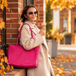 A woman in a beige coat and sunglasses stands outdoors by a brick wall holding an ice cream cone, carrying a large bright pink tote bag. Autumn leaves and trees are visible in the background.