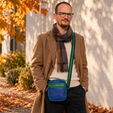 A man wearing a brown coat, cream sweater, and a brown plaid scarf stands outdoors against a white wall, wearing a blue crossbody bag with a green strap.