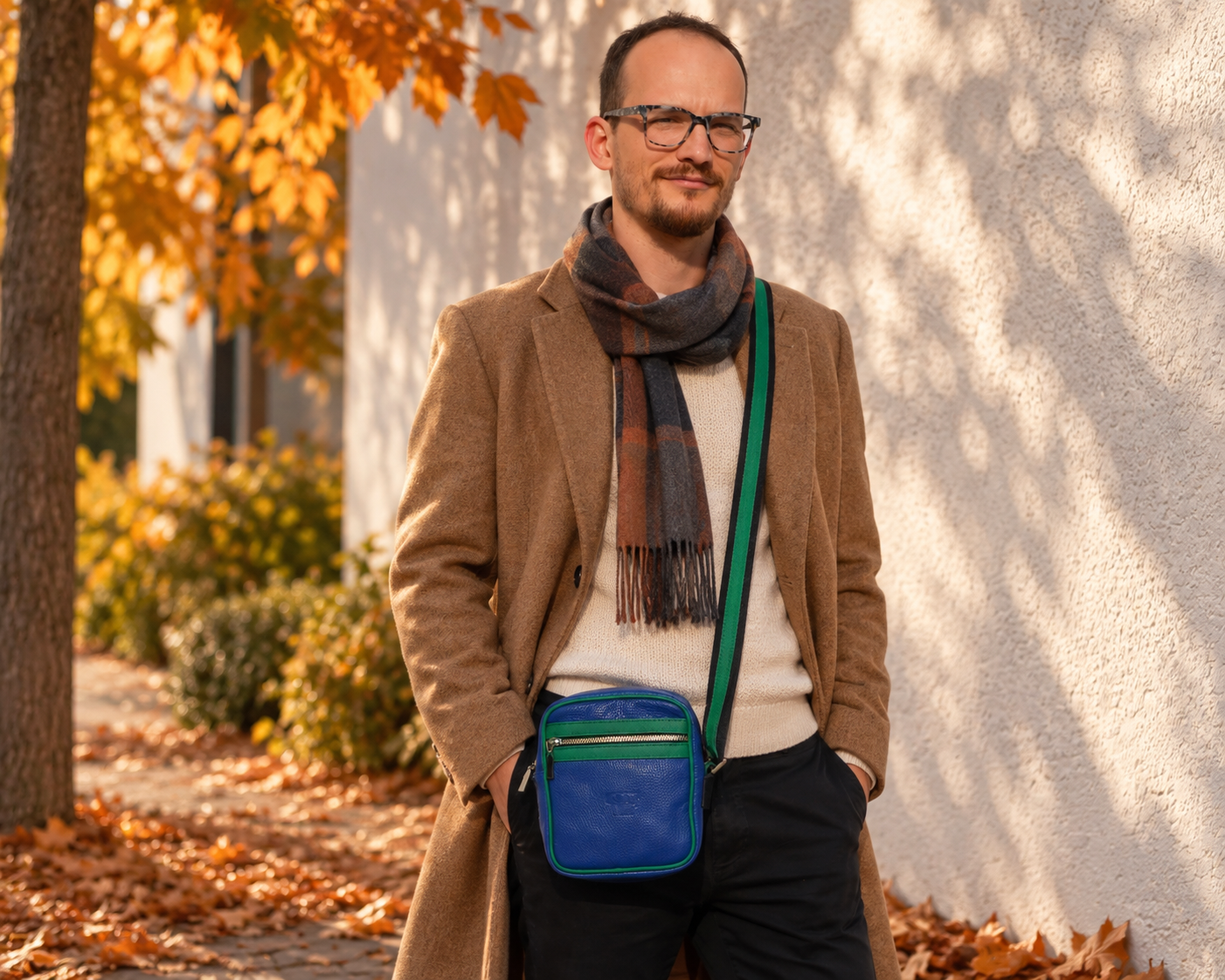 A man wearing a brown coat, cream sweater, and a brown plaid scarf stands outdoors against a white wall, wearing a blue crossbody bag with a green strap.