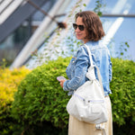 A woman in a light blue denim jacket and beige skirt stands outdoors in front of green bushes and a modern glass building, carrying a large white shoulder bag.