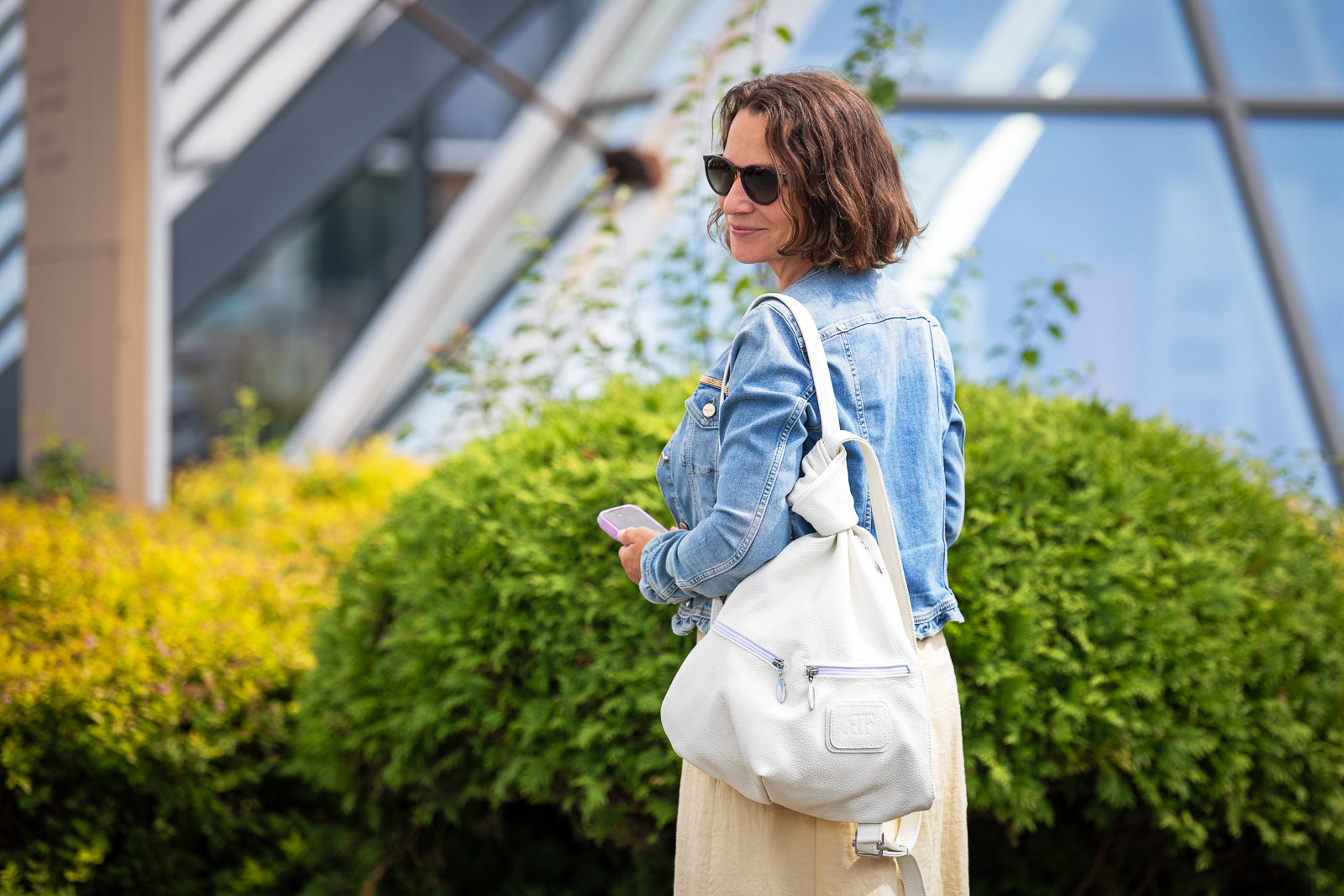 A woman in a light blue denim jacket and beige skirt stands outdoors in front of green bushes and a modern glass building, carrying a large white shoulder bag.