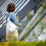 A woman viewed from behind wearing a light blue denim jacket and a cream-colored skirt, carrying a white canvas shoulder bag with a purple zipper pull. She is standing in front of a modern glass and metal structure with blurred green foliage in the foreground.
