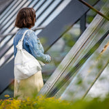 A woman viewed from behind wearing a light blue denim jacket and a cream-colored skirt, carrying a white canvas shoulder bag with a purple zipper pull. She is standing in front of a modern glass and metal structure with blurred green foliage in the foreground.