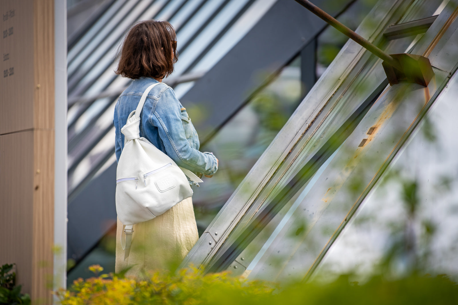 A woman viewed from behind wearing a light blue denim jacket and a cream-colored skirt, carrying a white canvas shoulder bag with a purple zipper pull. She is standing in front of a modern glass and metal structure with blurred green foliage in the foreground.