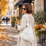 A woman in a beige trench coat and sunglasses stands on an autumn city sidewalk, carrying a large off-white shoulder bag. Fallen leaves cover the ground, and storefronts with potted orange flowers line the street.