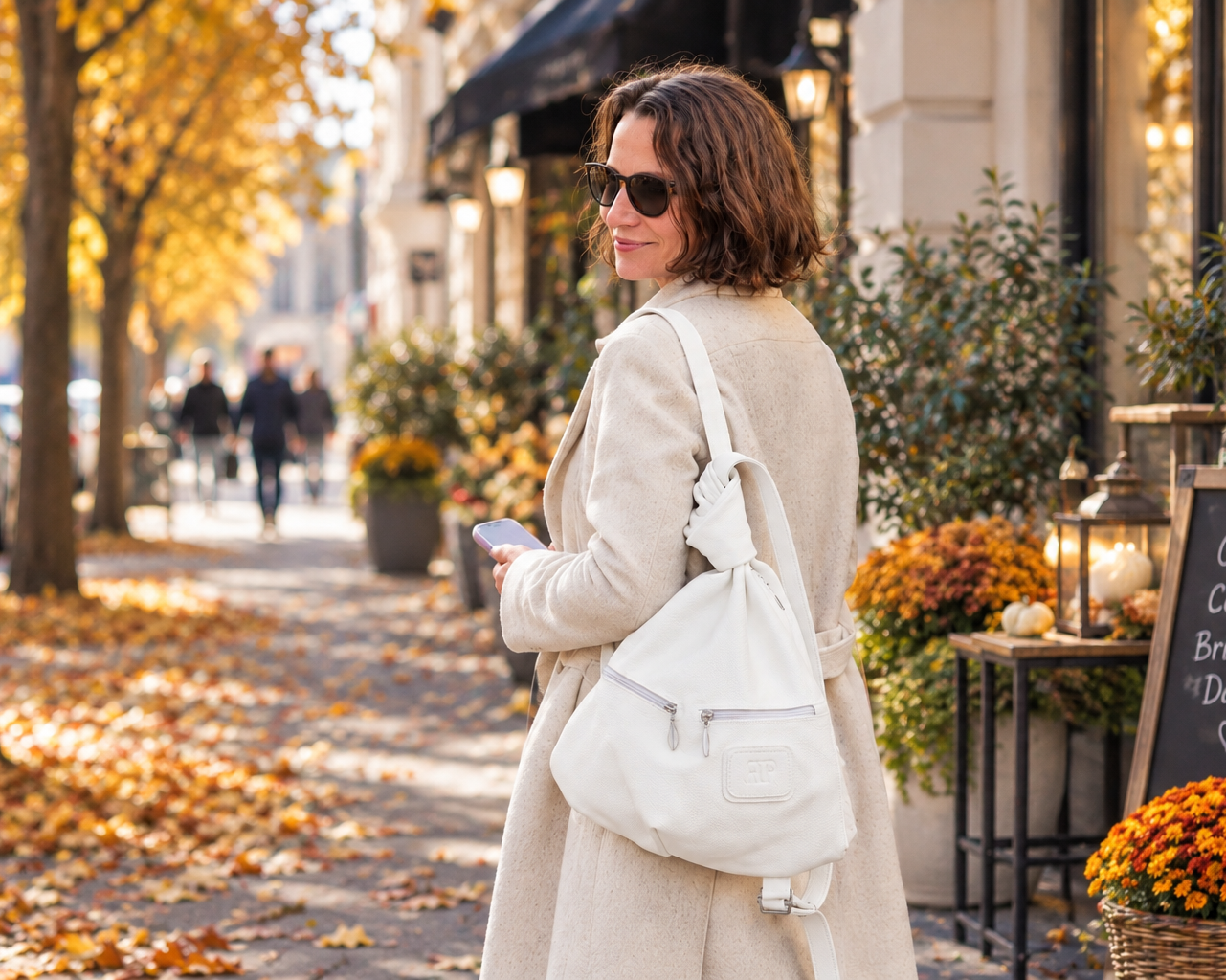A woman in a beige trench coat and sunglasses stands on an autumn city sidewalk, carrying a large off-white shoulder bag. Fallen leaves cover the ground, and storefronts with potted orange flowers line the street.